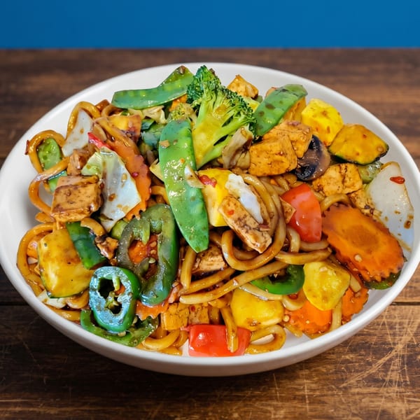 A white bowl filled with colorful stir-fried noodles, tofu, and mixed vegetables on a wooden table.