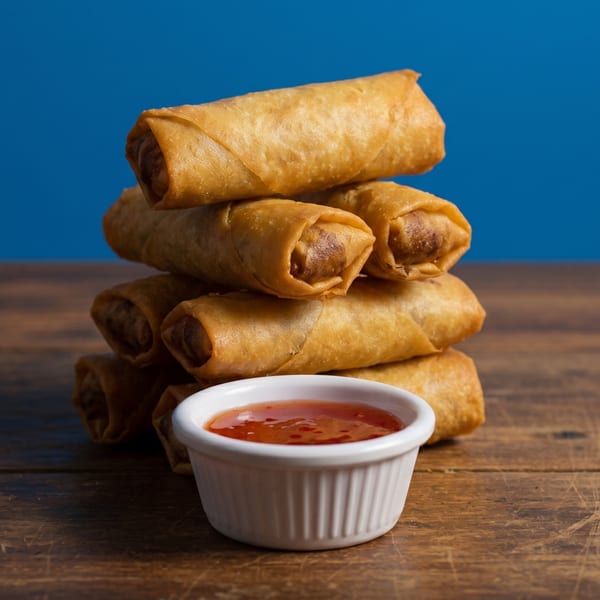 Stack of golden-brown egg rolls with a side of sweet chili dipping sauce on a wooden table.