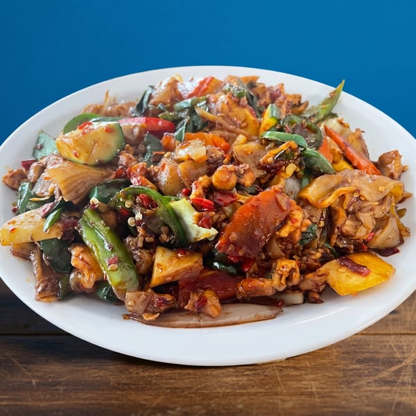 A white plate filled with spicy stir-fried noodles, vegetables, and meat on a wooden table.